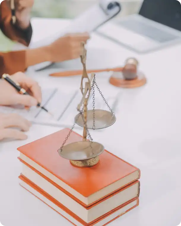 Wagi sprawiedliwości stoją na książkach, a w tle ludzie podpisują dokumenty przy stole.